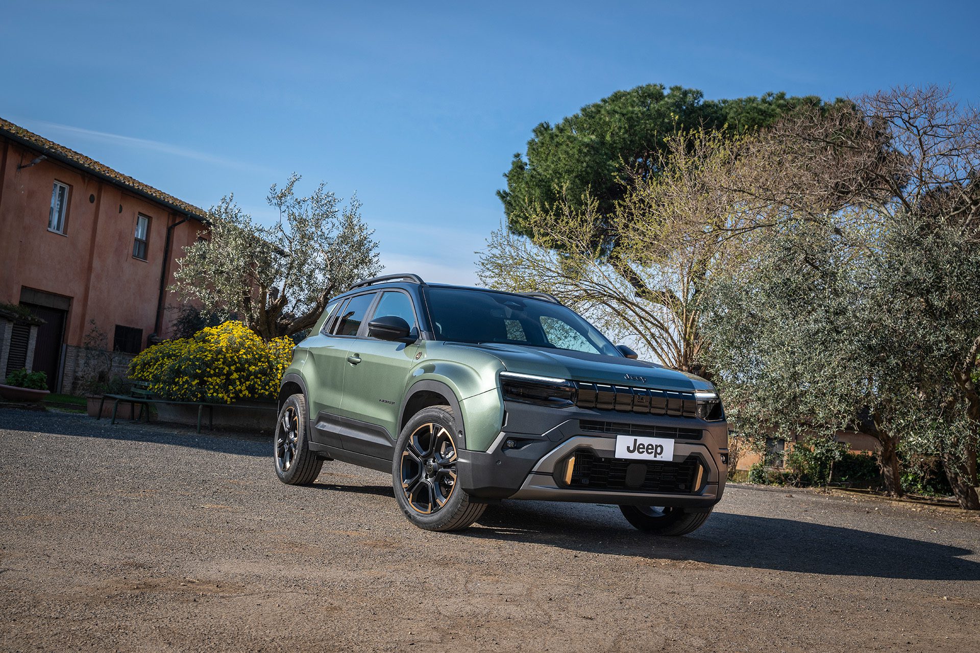 Groene Jeep Avenger 4xe schuin van voren bij bomen en een landelijk gebouw