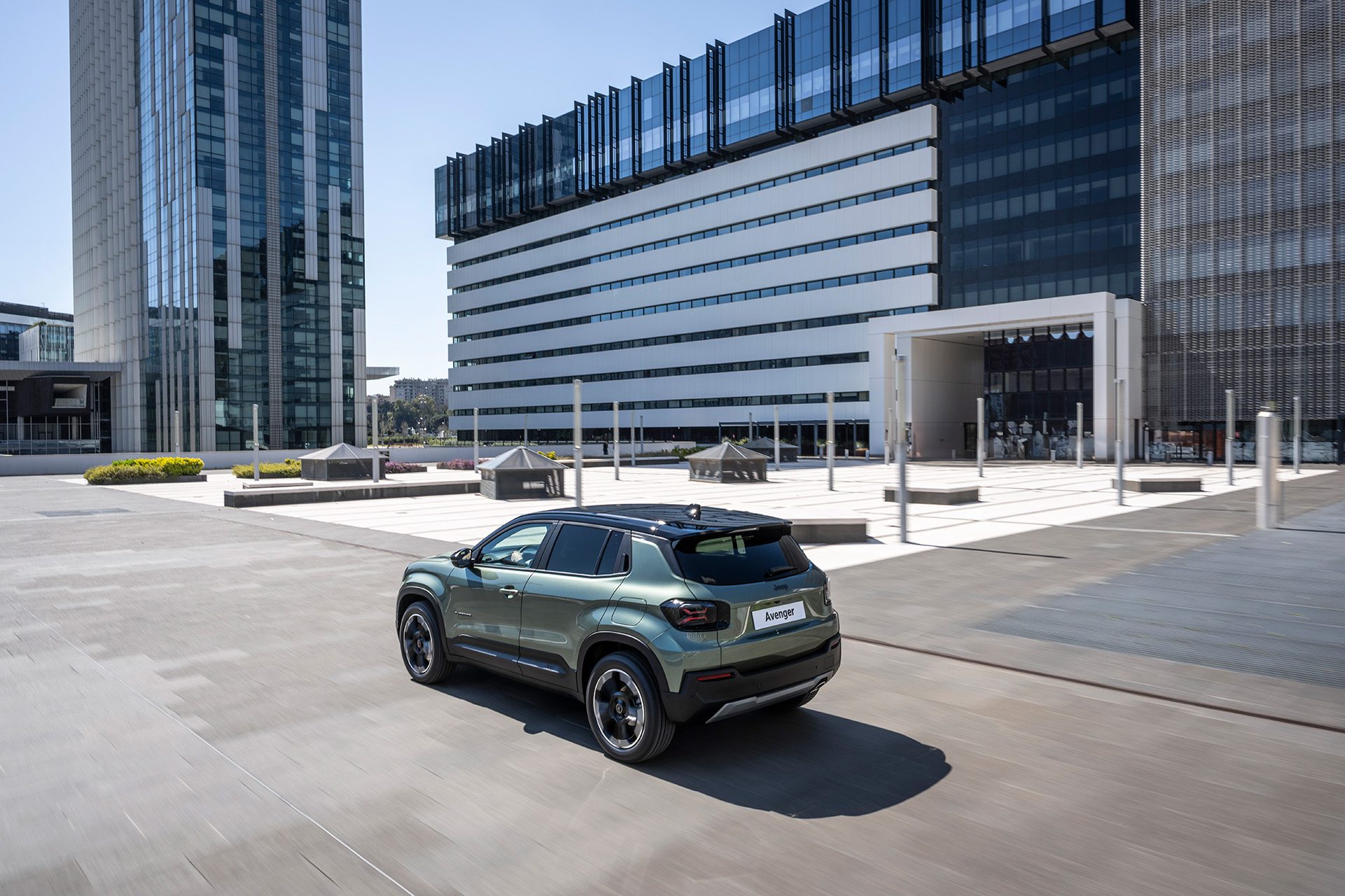 Groene Jeep Avenger Full Electric rijdt schuin van achteren door een moderne stadsomgeving